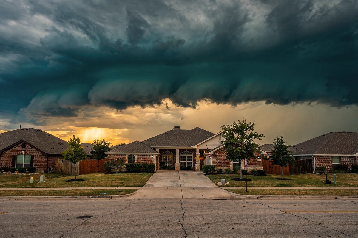 A dark shelf cloud spread above a straight subdivision street of brick houses with shingle roofs.